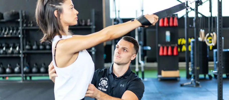 Kundin trainiert am Schlingentrainer, der Coach kniet daneben und führt die Bewegung